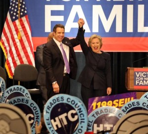 Hillary Clinton and Gov. Cuomo holding hands on a stage with people holding signs saying HTC in front of them