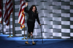 Tammy Duckworth walking on stage with her prosthetic legs, wearing a black suit with American flag in background