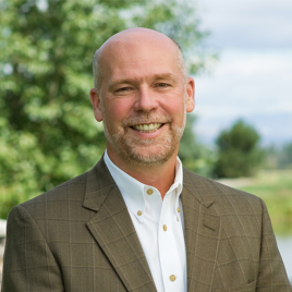 headshot of Greg Gianforte wearing a white collared shirt and brown blazer, outside with a tree in the background