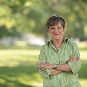 Laura Kelly standing and smiling with arms crossed wearing a green shirt in front of a blurred out green tree background