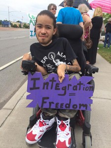 A young African American man, using a power wheelchair, holds a purple sign that reads "Integration = Freedom"