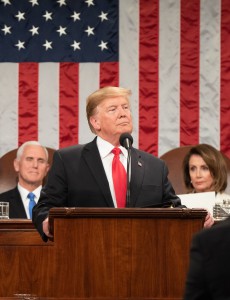 President Trump standing behind the podium of the House of Representatives, with Vice President, Mike Pence (on the left) and the Speaker of the House, Nancy Pelosi (on the right) seated behind him. Behind all three hangs the United States Flag. 