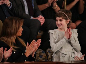 Image Description: Grace Eline, a 10-year-old girl and cancer survivor, is dressed in a ruffled gray, long-sleeved dress, with a pixie haircut and studded headband. She is standing to accept President Trump's recognition of her, while clapping her hands. The First Lady, Melania Trump, who is seated next to her, smiles in her direction, also clapping. 