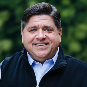 Illinois Governor JB Pritzker smiling in front of a blurred background of green