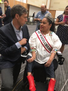 Beto O'Rourke is crouched on his knees talking a latina woman with a Ms. Wheelchair sash and tiara in a power wheelchair.