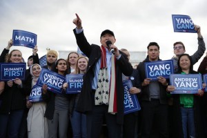 Yang talks into a microphone with his arm outstretched  with fans behind him holding signs