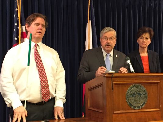 Image of Jeff James in a white shirt and red tie holding a white cane standing next to Gov. Terry Branstad and Lt. Gov Kim Reynolds who are standing behind a podium.