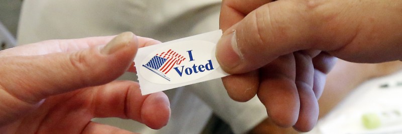 Image of a male hand handing an "I Voted" sticker to another hand