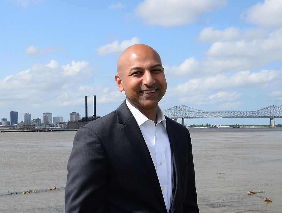 Abhay Patel standing and smiling facing the camera wearing a white dress shirt and black blazer with clouds, a bridge and water in the background