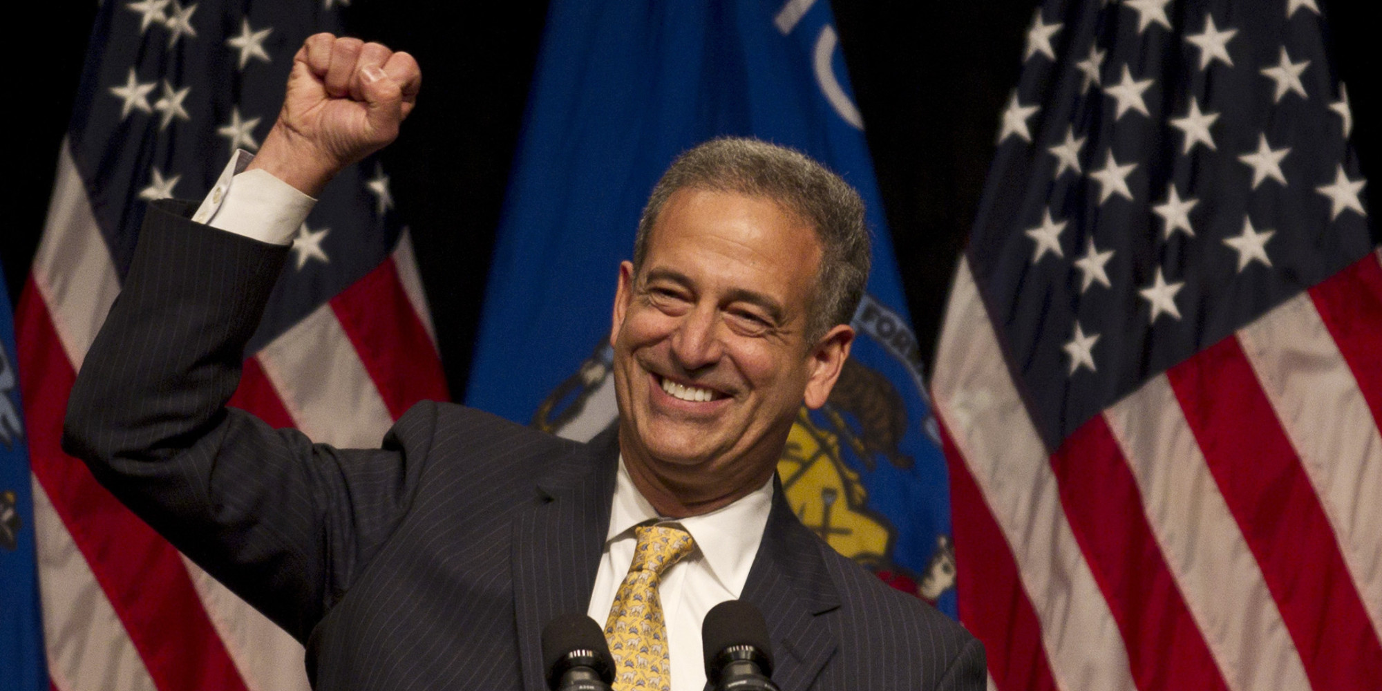 Russ Feingold smiling with a fist in the air while speaking at a campaign event. American flags in the background