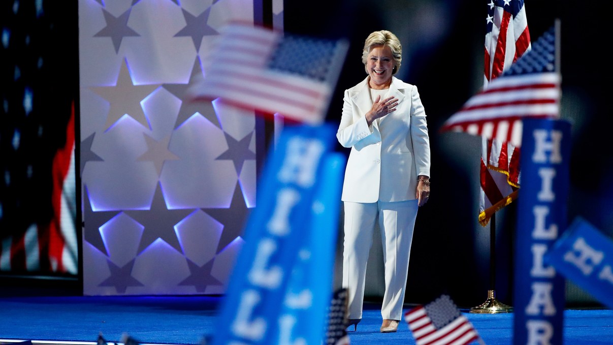Hillary Clinton standing on stage with her hand on her heart, with Hillary signs and American flags waving in foreground
