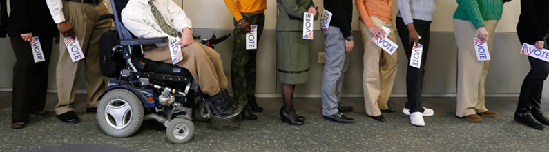 Image shows people in line waiting to vote. Majority are physically-able-bodied individuals standing in line. One man is in a power wheelchair.