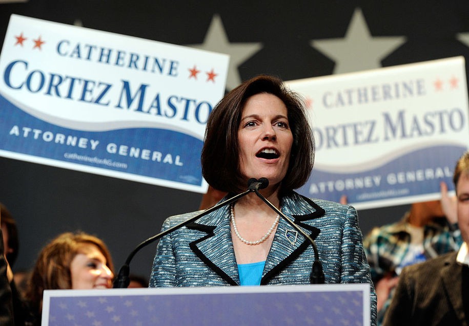 Catherine Cortez Masto speaking at a rally wearing a blue suit with signs saying her name behind her