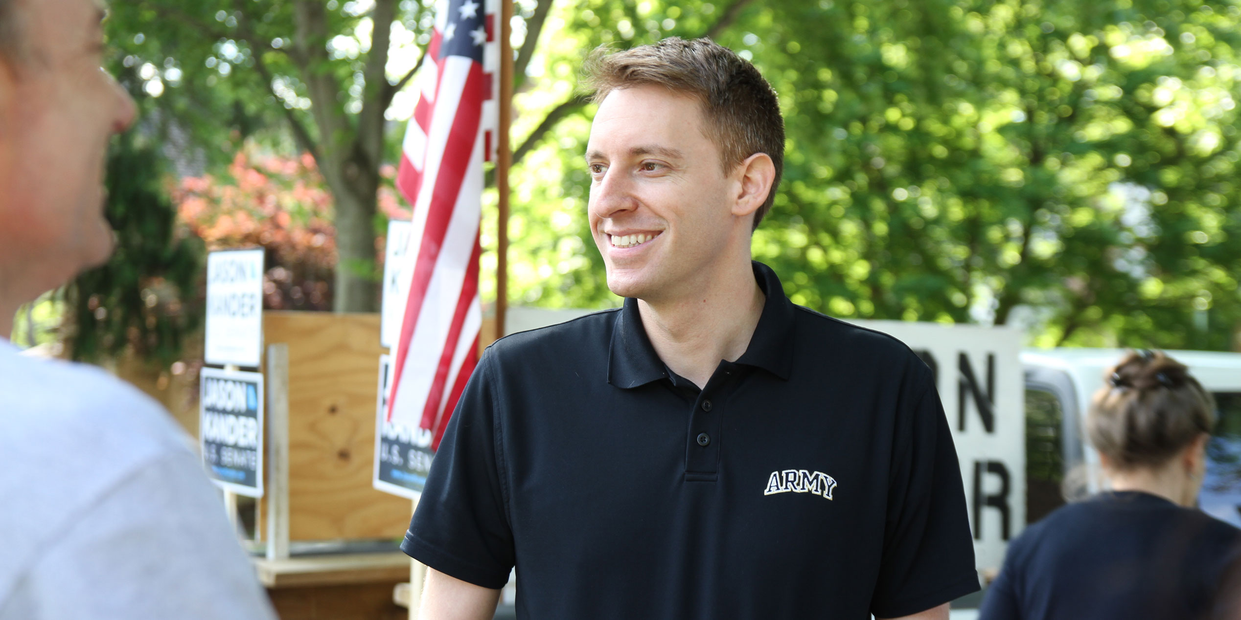 Jason Kander wearing an Army shirt standing outside with an American flag in the background