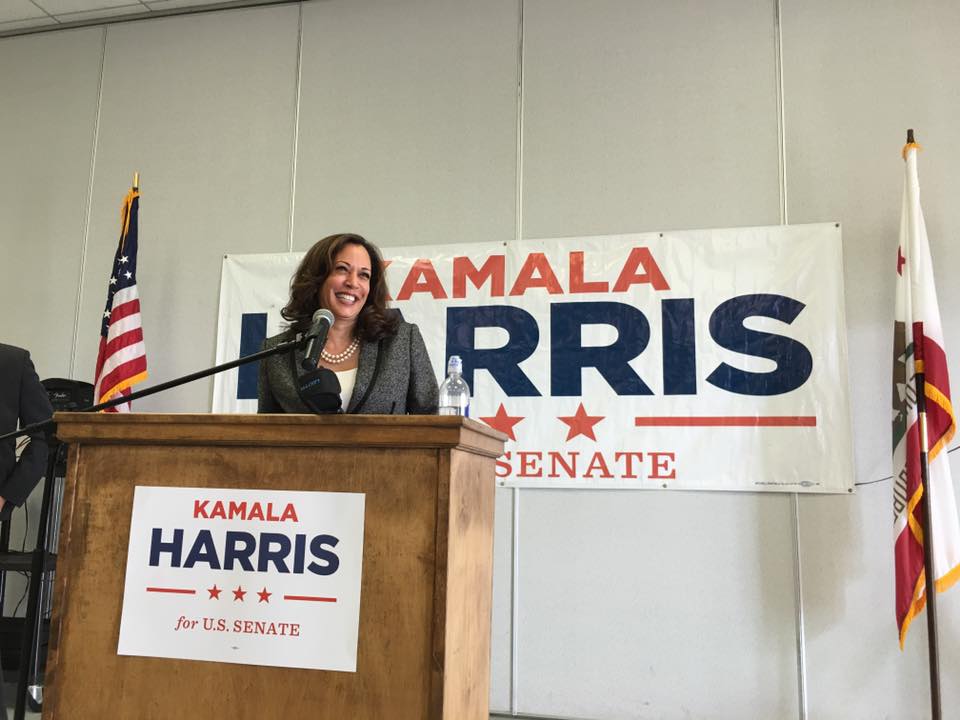 Kamala Harris speaking behind a podium and microphone with sign saying her name in the background