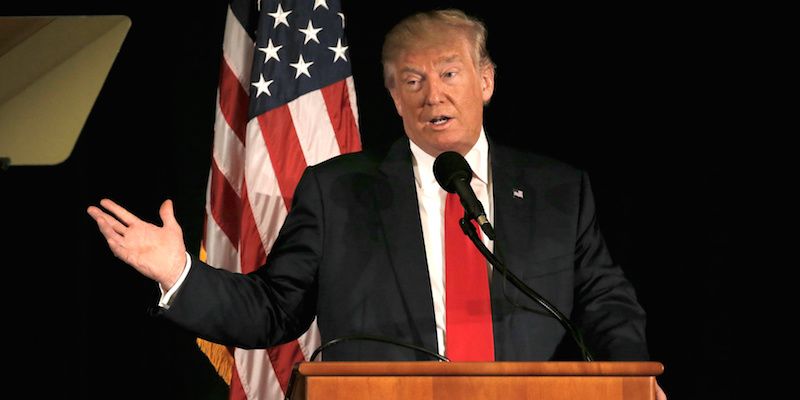 Donald Trump wearing a black suit, white shirt and red tie speaking behind a podium with a microphone and an American flag in background