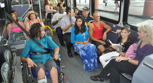 A young African American woman in a wheelchair is strapped in on a bus with a diverse group of people seated on the bus around her.