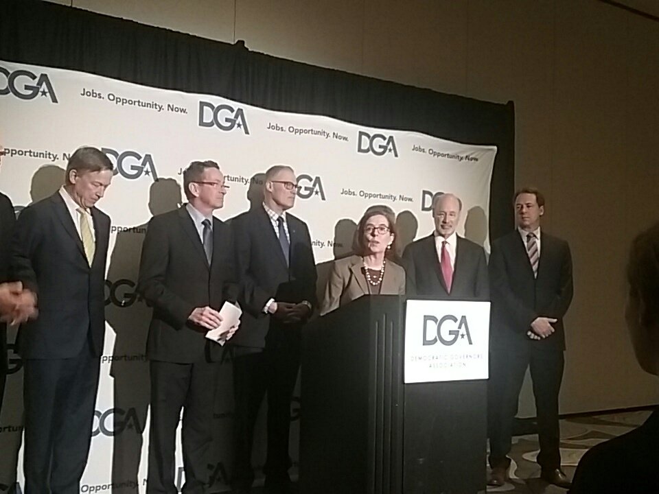 Gov. Brown speaking at a podium with a sign saying DGA surrounded by other Democratic governors standing behind her