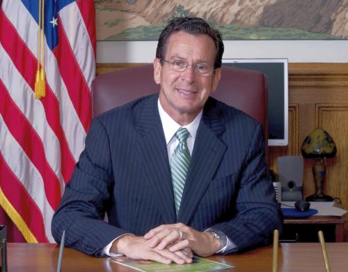Gov. Dannel Malloy wearing a suit seated behind a large wooden desk with an American flag in the background