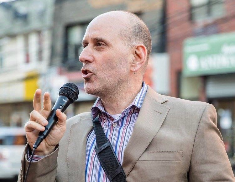 Harvey Epstein speaking into a microphone in front of a blurred background of storefronts in New York City