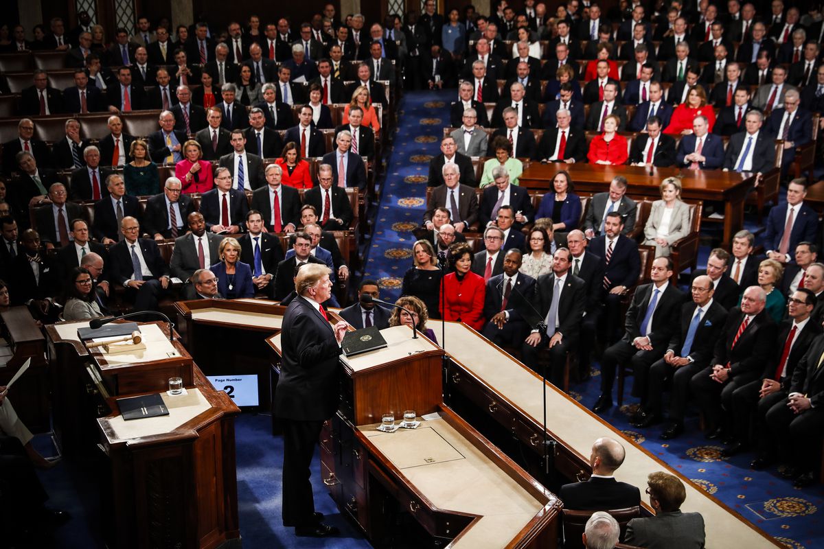 President Trump is standing behind the podium of the House of Representatives delivering his State of the Union Address, looking into a crowd of seated legislators in the auditorium.