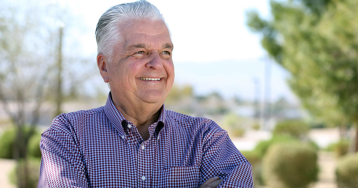 Steve Sisolak smiles in front of a blurred background of trees and mountains