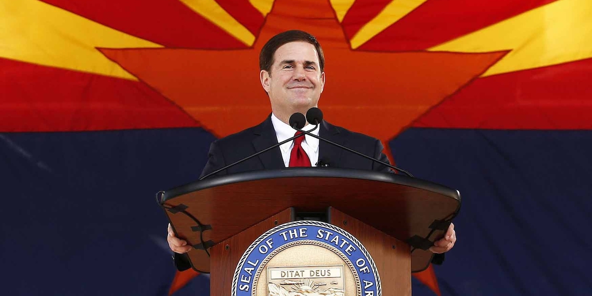 Arizona Governor Doug Ducey smiling behind a podium in front of the Arizona state flag