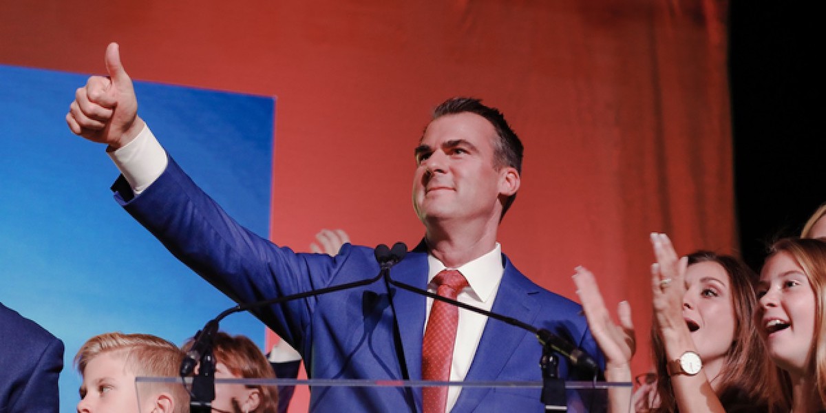 Governor Kevin Stitt giving a thumbs up on stage with supporters behind him