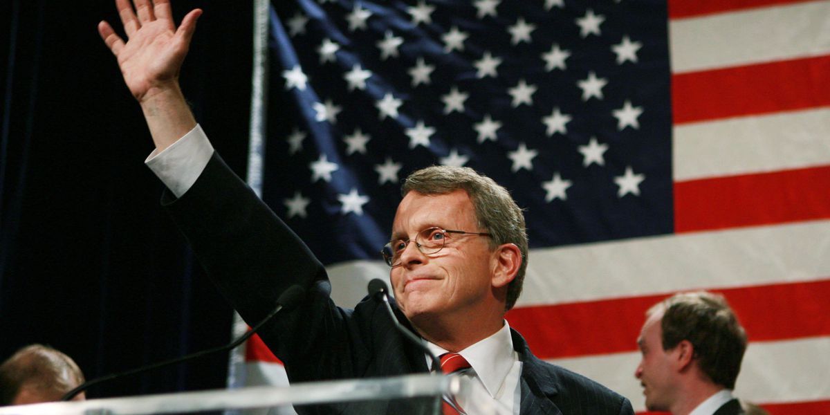 Governor Mike DeWine with his right hand raised in the air in front of an American flag on stage