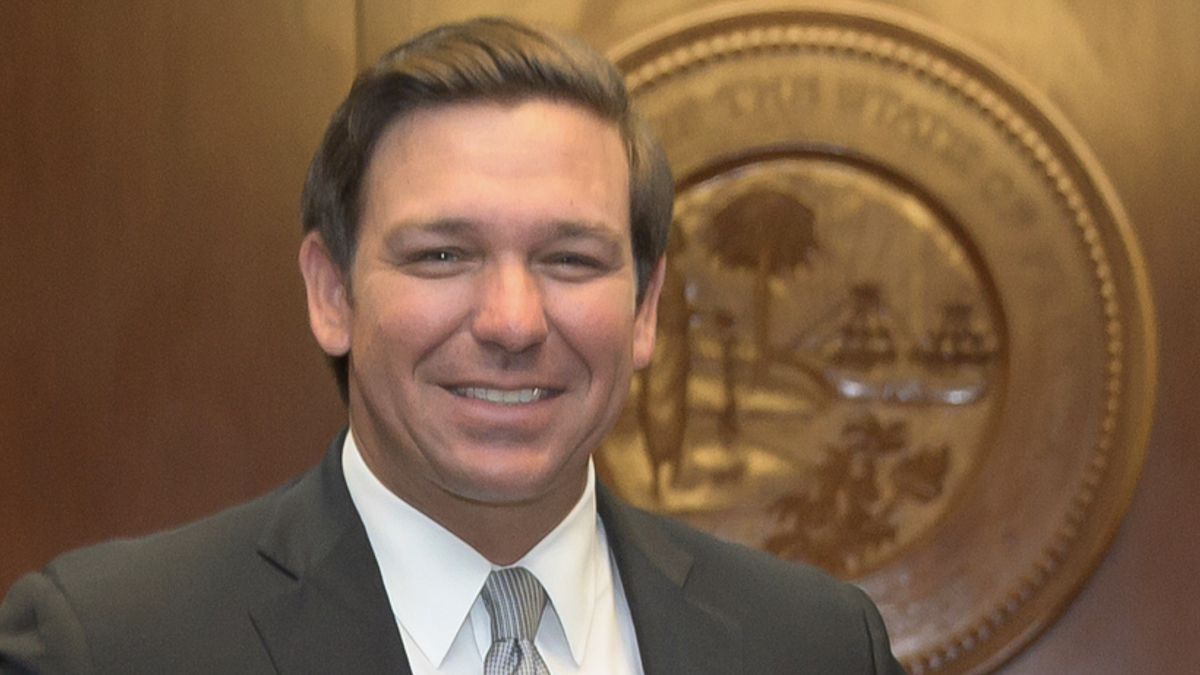 Ron Desantis smiles in front of a sign for the state of Florida