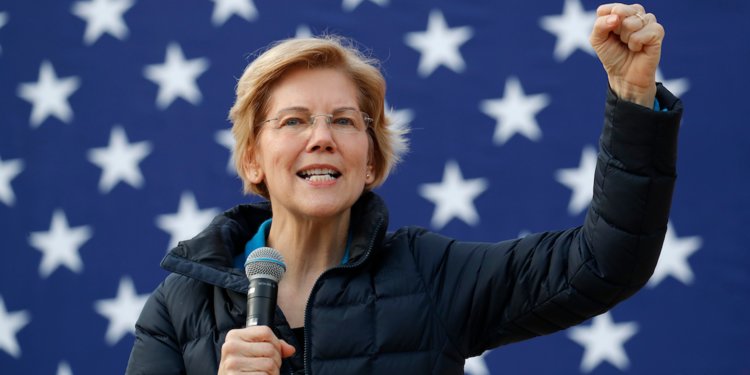 Warren speaks into a microphone with her arm outstretched in front of the U.S. flag