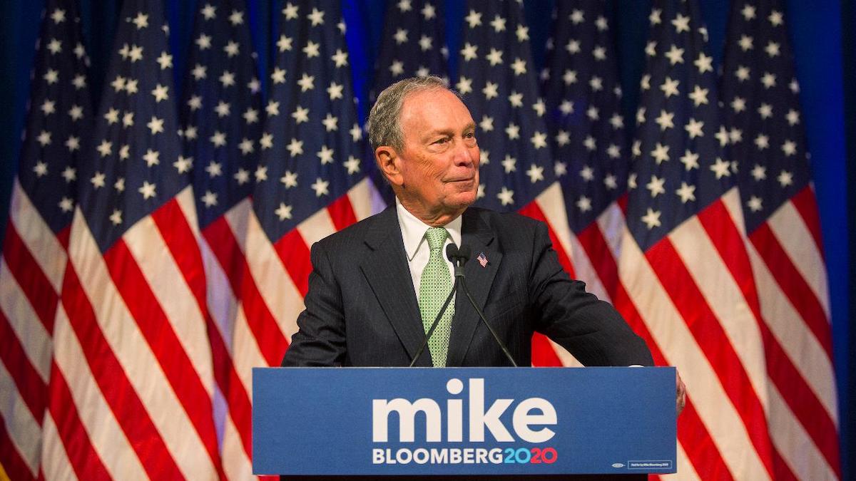 Michael Bloomberg speaking at a podium in front of American flags. The podium has his campaign logo on it.