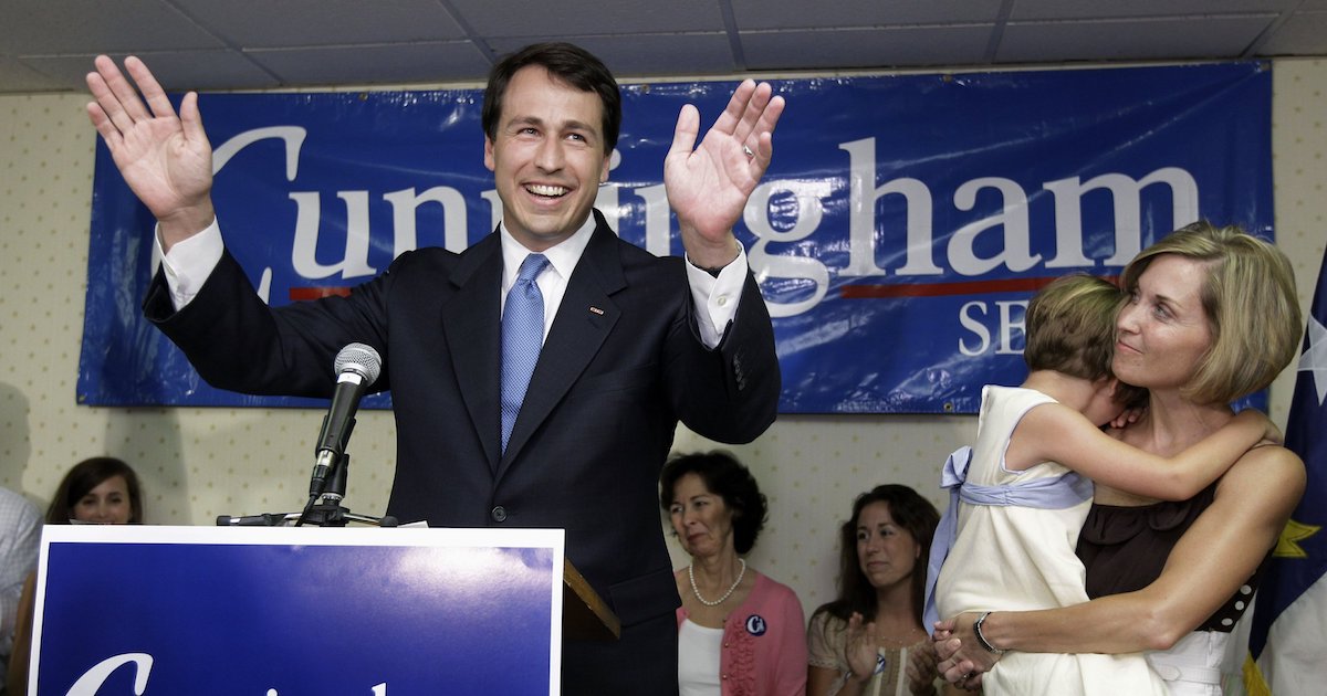 Cal Cunningham speaking at a campaign event in front of a sign with his logo on it, next to a woman holding a child