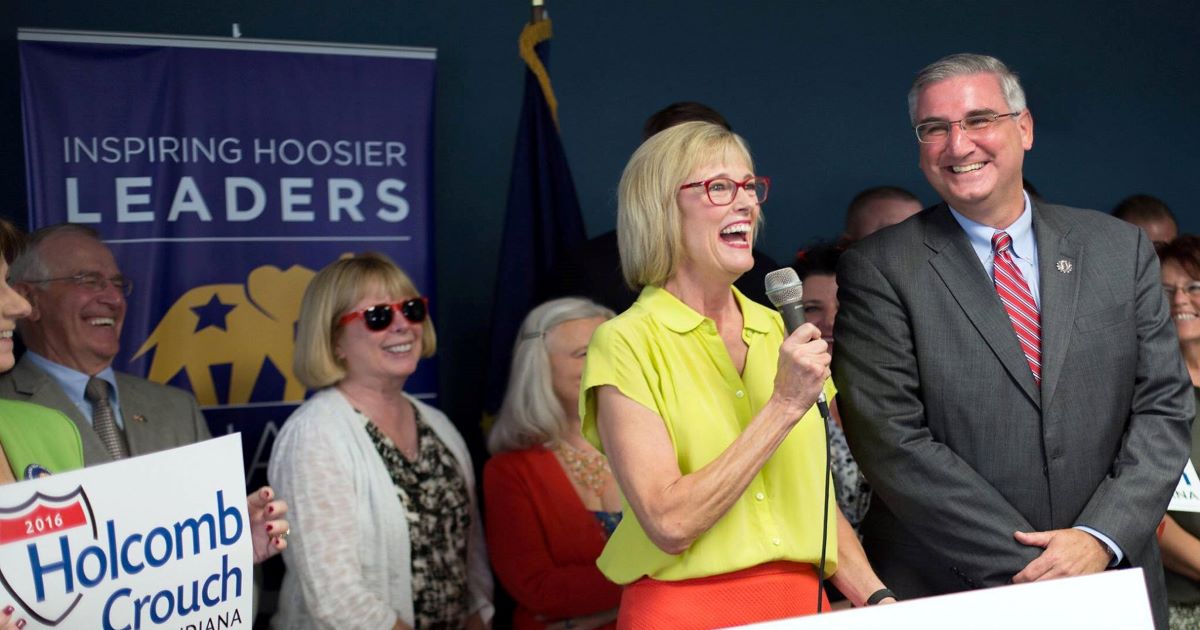 Eric Holcomb and Suzanne Crouch stand behind a podium with supporters behind them