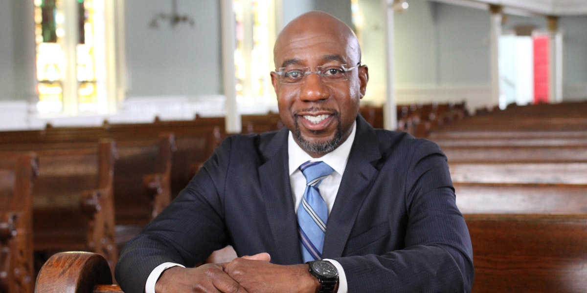 Reverend Raphael Warnock smiling wearing a suit and tie in an empty church